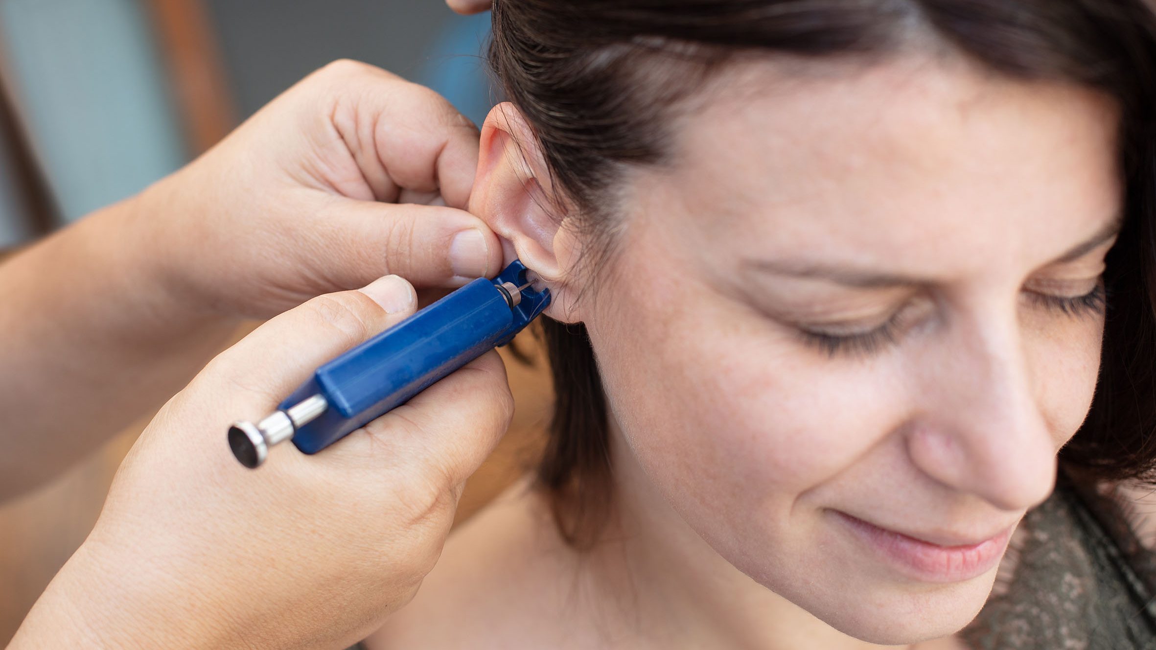 Woman having ear piercing process with special piercing gun in beauty center by medical worker, cropped close up view